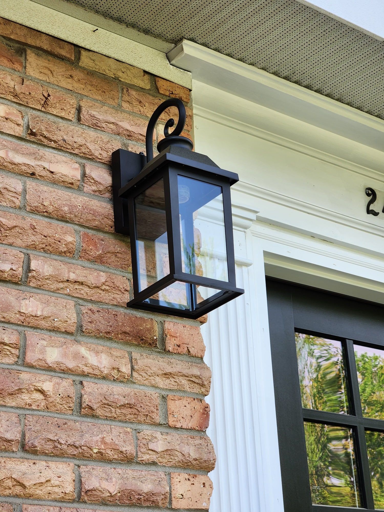 A black square-lantern wall light mounted on red brick beside a white-trimmed window, its scrolled arm bracket fixed to the mortar line under the soffit.