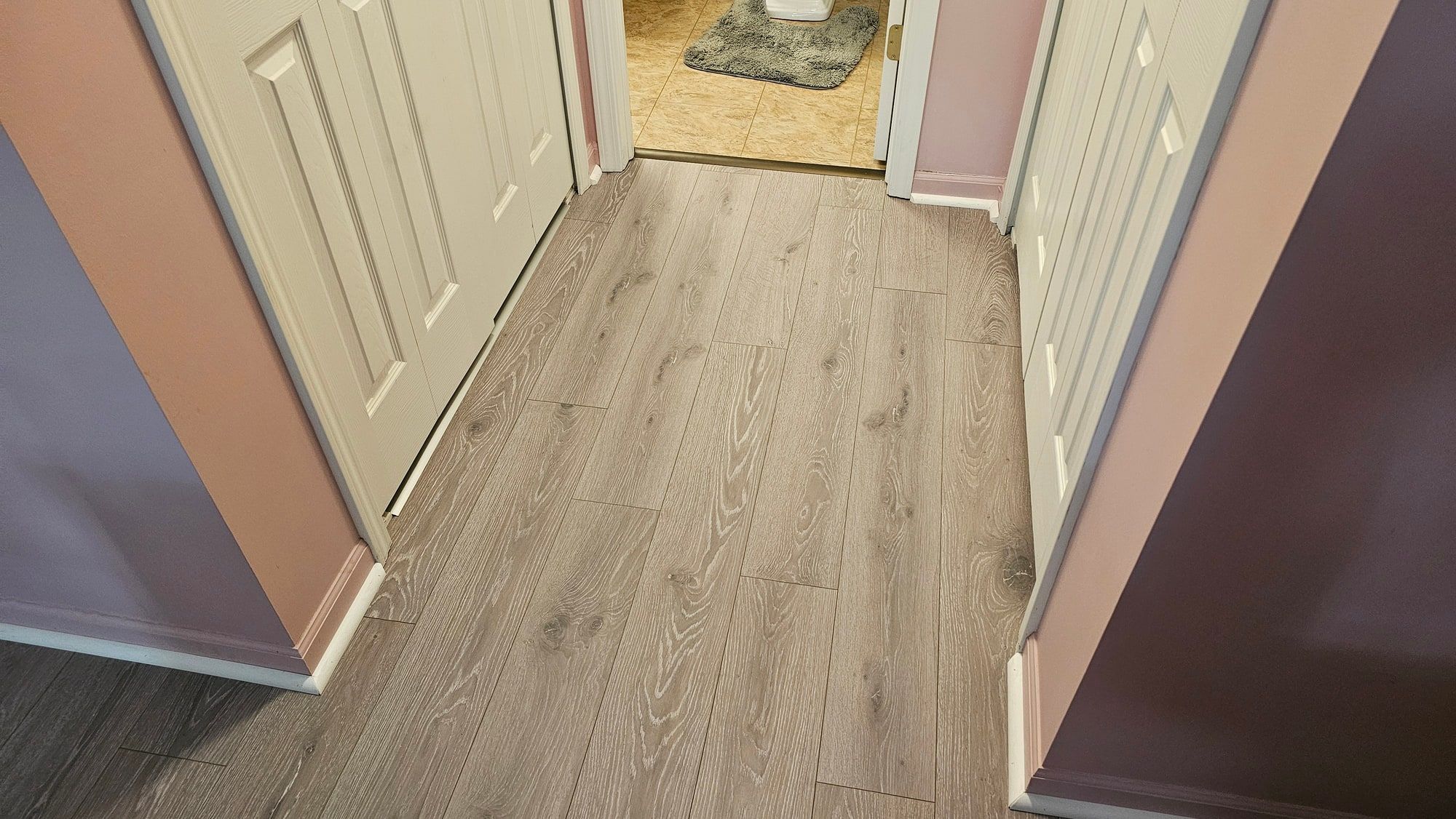The same hallway in light oak-look plank running the length of the hall, white baseboard back on, a threshold strip at the bathroom tile.
