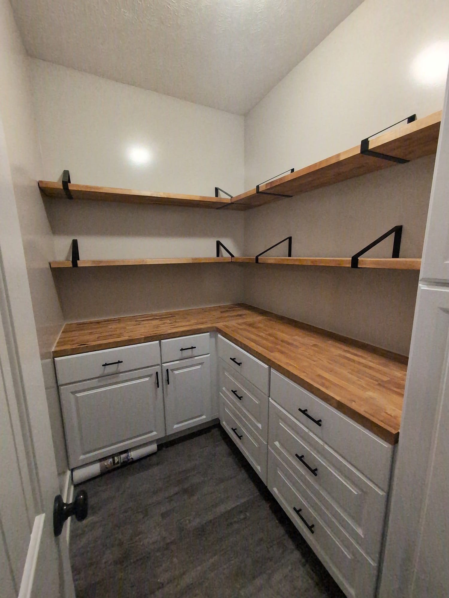 The same pantry with a butcher block counter along two walls over white shaker cabinets and drawers, and butcher block shelves above on black brackets.