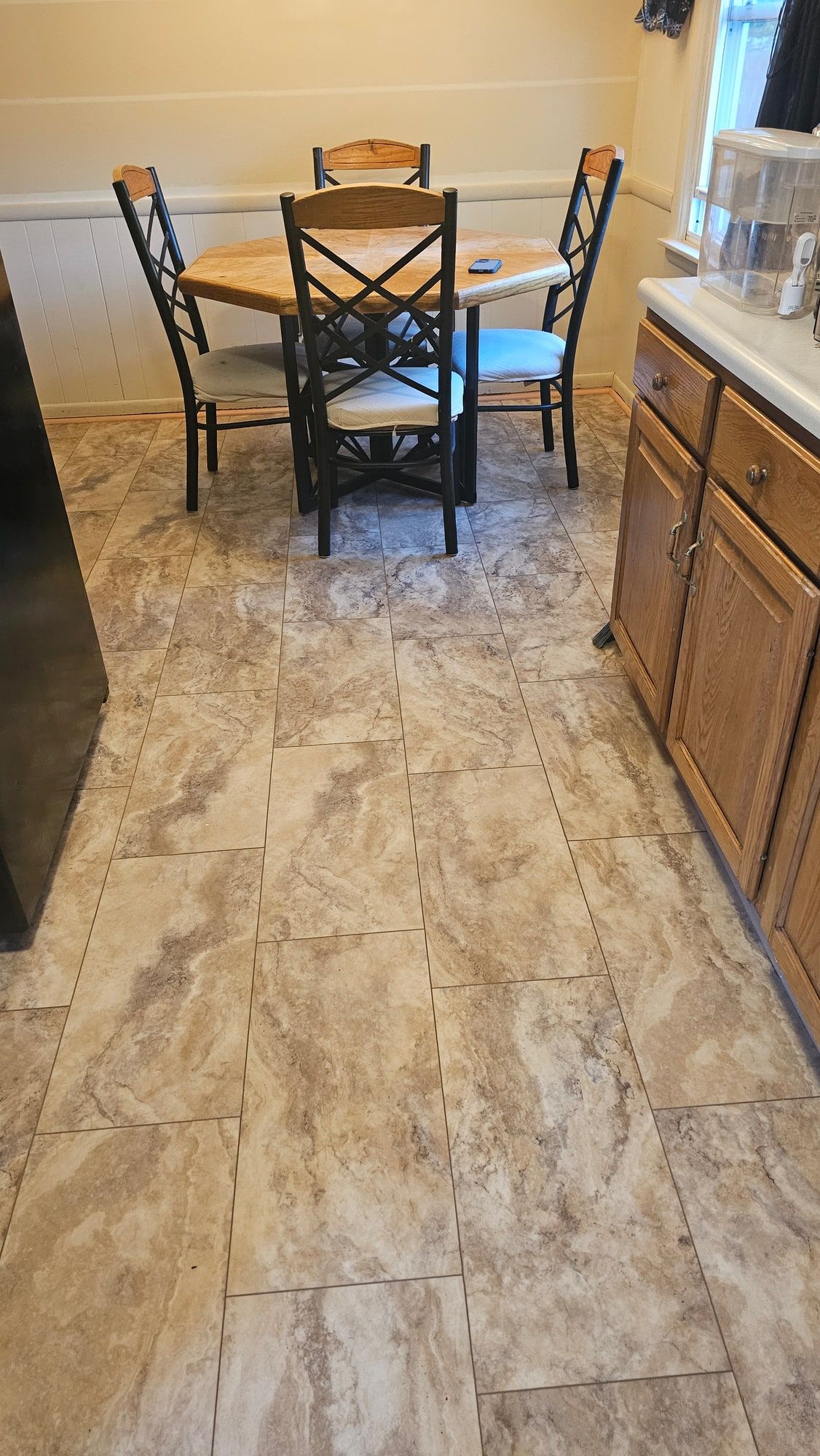 The same kitchen floor in large stone-look tile laid in a running bond, carried under the table and up to the cabinet toe kick.