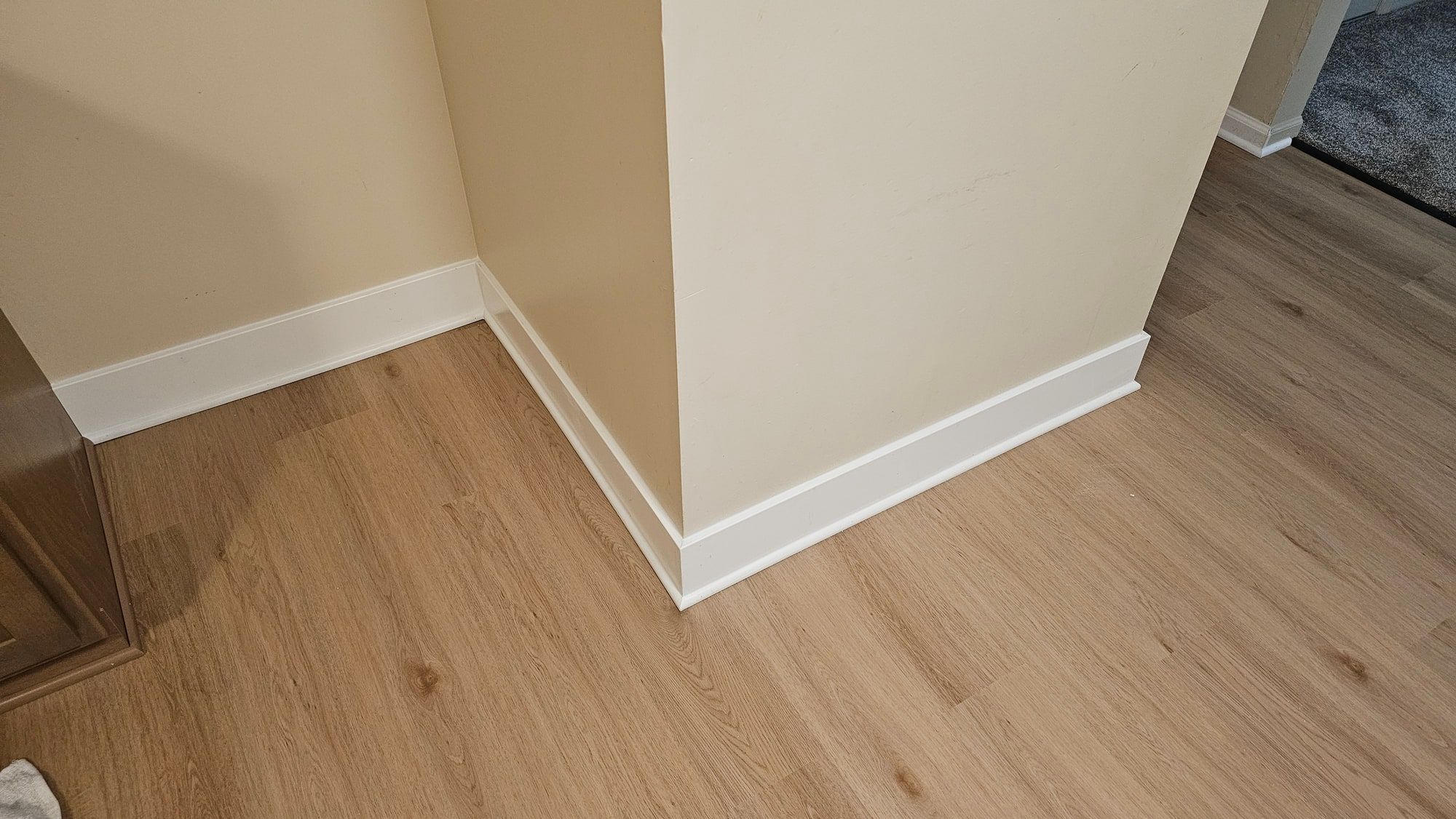 An outside corner where two runs of white baseboard meet over light oak-look plank flooring, the miter closed tight, the plank scribed around the door casing.